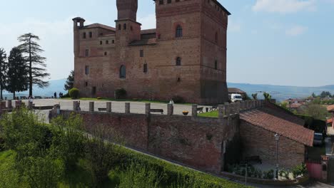 Dolly backward shot of Grinzane Cavour castle, revealing its historic brick architecture, manicured gardens, and the surrounding Langhe hills under a serene blue sky