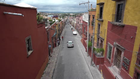 a street in san miguel de allende, guanajuato mexico with yellow and red houses, some cars and people