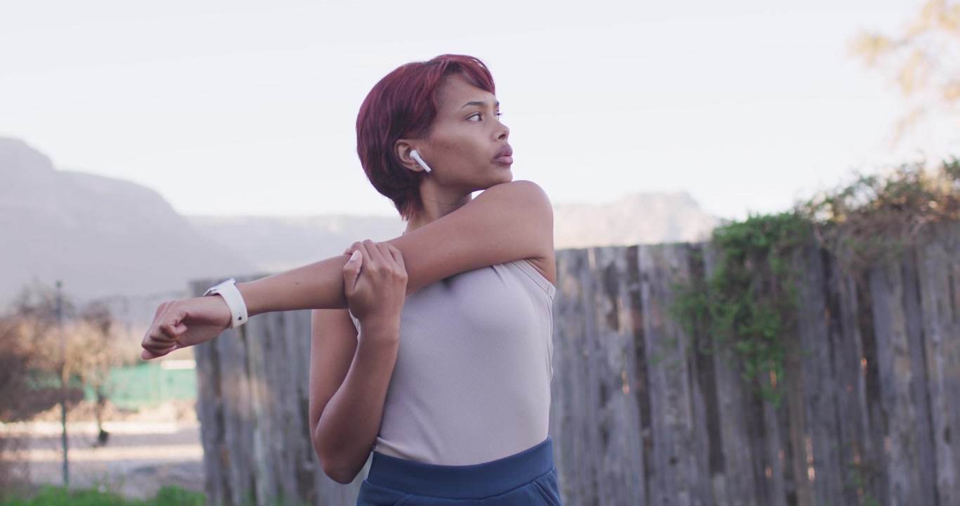 African American woman doing crossbody stretches warming up for run by wooden fence with smartwatch