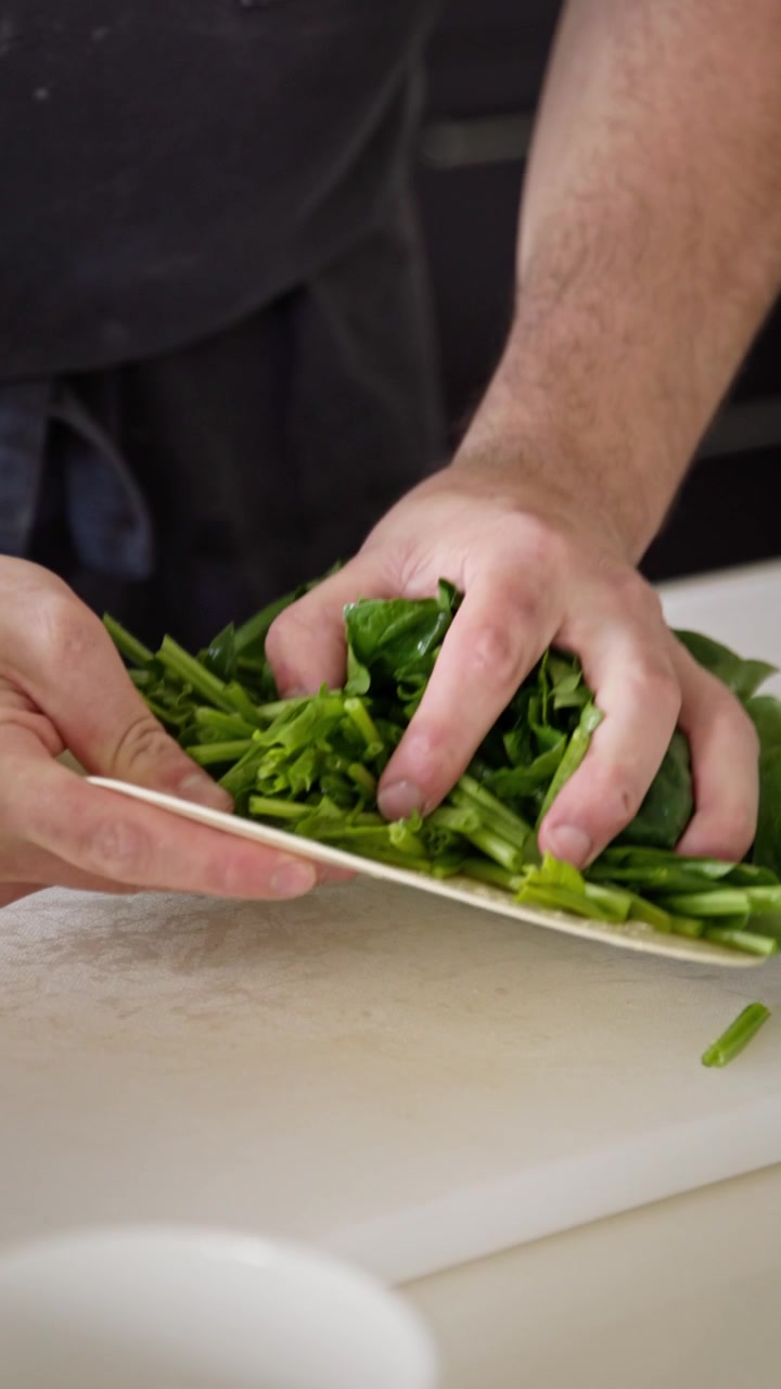 Primo piano di mani che trasferiscono spinaci tritati in una ciotola di metallo usando una spatola. Scatto verticale di preparazione in cucina che mostra verdure fresche aggiunte durante la preparazione del cibo.