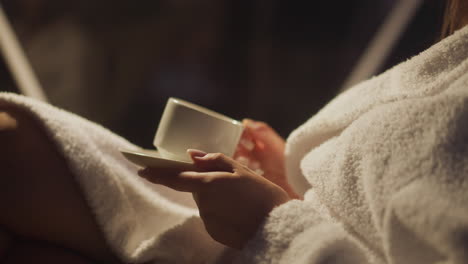 resting woman in bathrobe drinks hot beverage in cup enjoying being alone. calm and quiet atmosphere in semi lit hotel room. peace of mind alone with thoughts