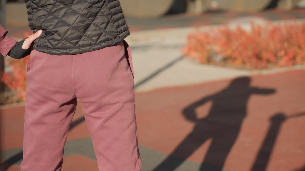 Back view of student workout participant swaying waist sideways on sunny outdoor court with autumn shrubs behind, long shadows of figure stretching across red gym mats, focus on form and movement