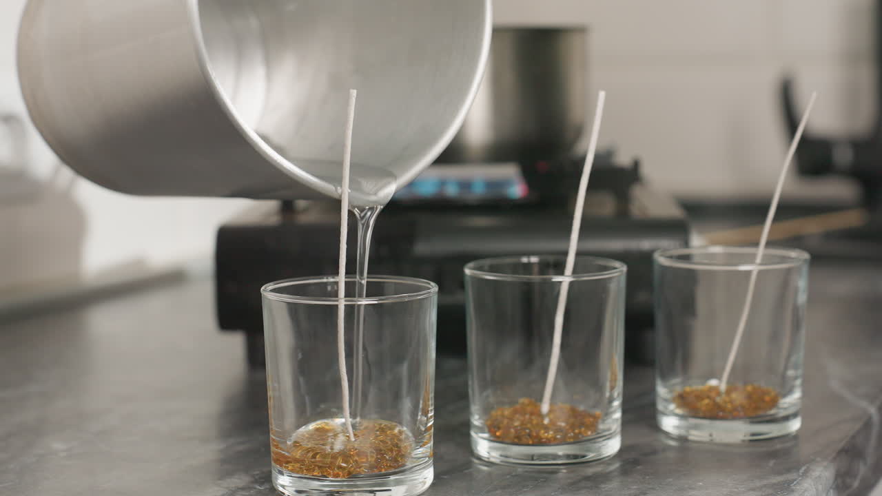 Candle wax production process shown as person carefully pours melted wax from metal jug into three aligned glass cups with centered wicks on kitchen counter with pot heating on gas burner