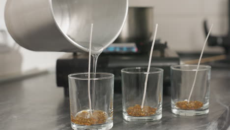 candle wax production process shown as person carefully pours melted wax from metal jug into three aligned glass cups with centered wicks on kitchen counter with pot heating on gas burner