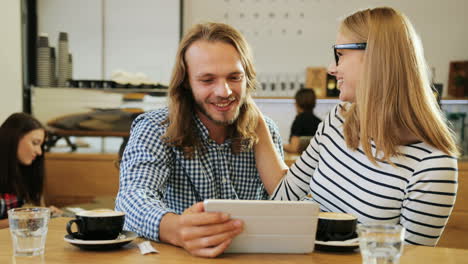 caucasian happy friends laughing and watching a video on a tablet sitting at a table in a cafe