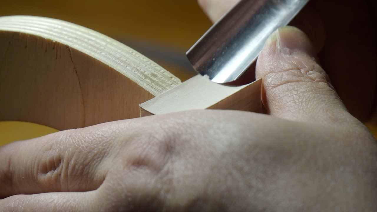 Geigenbauer verfeinert Zarge und Kante einer neuen Geige auf einer Innenform mit einem kleinen Geigenbauer-Hobel. Vorsichtiges Abhobeln des Tonholzes zur Definition des Korpusumrisses mit präzisen, kontrollierten Zügen. Extreme Nahaufnahme.