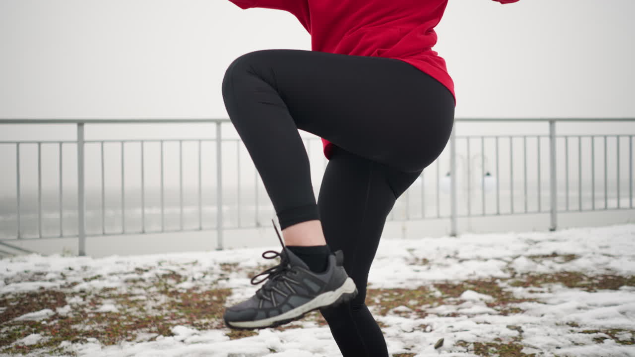 야외에서 운동하는 여성, 눈 인 땅에서 운동하는 동안 다리를 들어 올리고, 뒷면에 흐릿한 조명 기둥이 있는 철의 레일링 근처에, 은 후디와 검은 운동복을 입고 따뜻함을 얻습니다.
