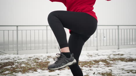 mujer haciendo ejercicio al aire libre, levantando la pierna durante el entrenamiento en un terreno nevado cerca de una barandilla de hierro con un poste de luz borroso en el fondo, usando una sudadera roja con capucha y ropa deportiva negra para el calor