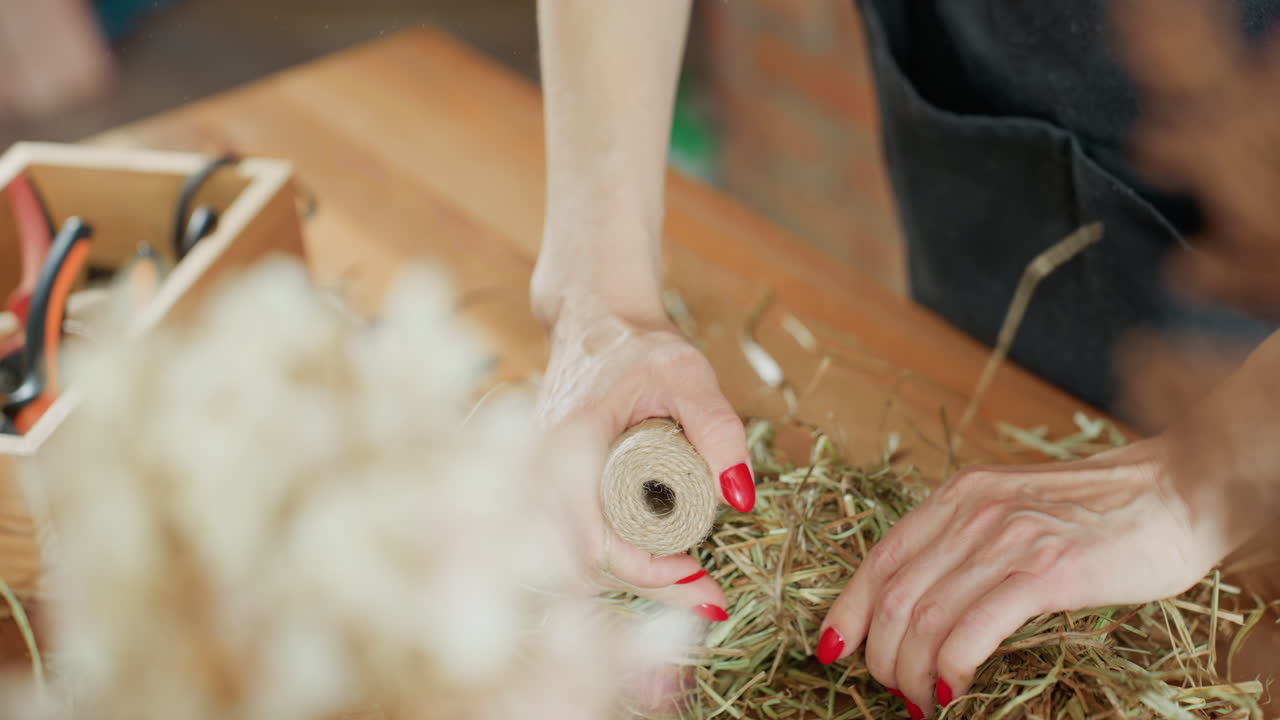 Decorator arranging hay by hand on circular base for natural wreath creation, red nails visible while working on rustic handmade decoration design project using organic materials for seasonal craft process