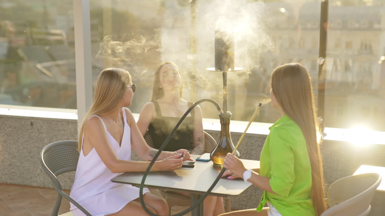 Company of young females sitting at the table on the roof of the building. Girls talking, smiling and smoking hookah.