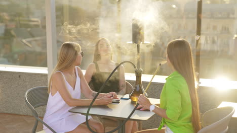 company of young females sitting at the table on the roof of the building. girls talking, smiling and smoking hookah.