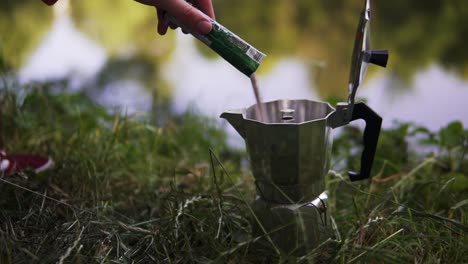 verter café en polvo dentro de una lata de café plateada retro para tomar una copa en el desayuno en un viaje de campamento junto a un lago o río