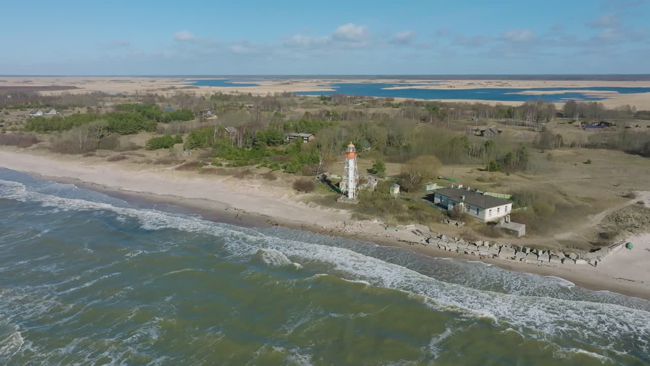 vista aérea do farol de cor branca, costa do mar báltico, letónia, praia de areia branca, grandes ondas a quebrar, dia ensolarado com nuvens, foto de ampla órbita de drone