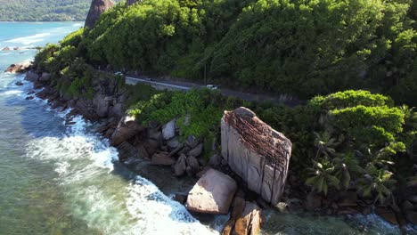side drone shot of passing cars on road near cliff, huge granite rock at anse forbans beach mahe seychelles