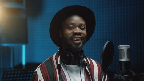 portrait of the handsome young guy in a hat standing at the mic in the dark sound studio, looking and smiling to the camera