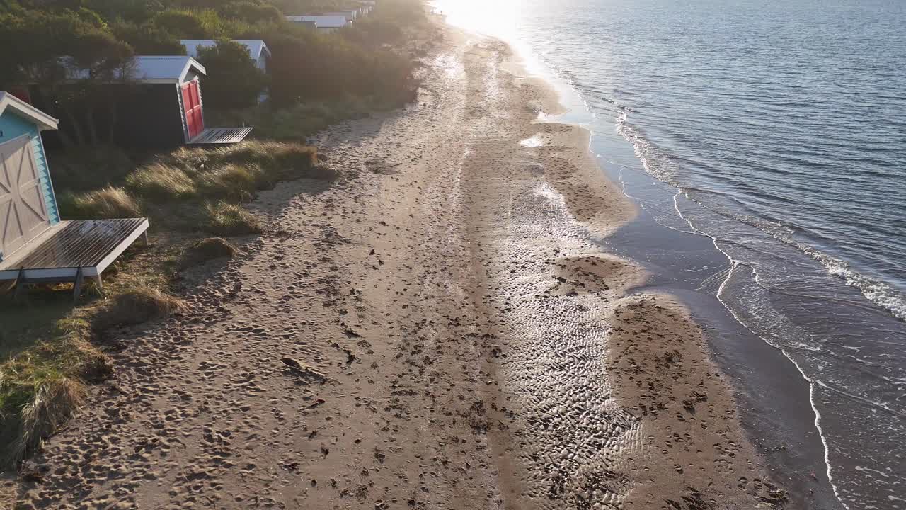 Drone glides above sandy shoreline, colorful beach huts, and calm sea in golden morning light