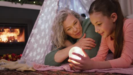 Happy-caucasian-girl-with-grandmother-playing-with-snow-globe-at-christmas