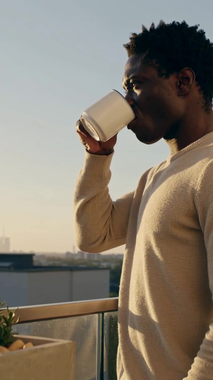 Vertical video: Sunlight drawing man in light sweater sipping white mug on balcony