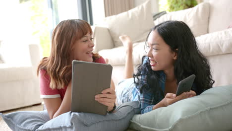 two biracial young female friends relaxing at home, using a tablet and a smartphone