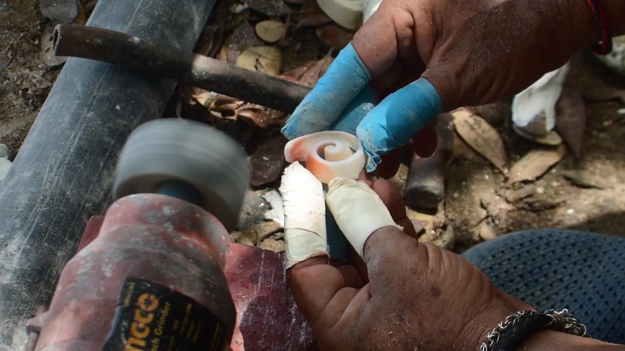 Artisan polishing natural seashell against high-speed rotary buffing wheel during final finishing stage of handcrafted production. Fine shell dust is visible in the air as the rotating wheel smooths