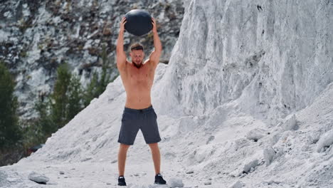 Concentrated muscular man exercising with black ball in the mountains. Strong shirtless guy doing squatting with heavy ball outdoors.