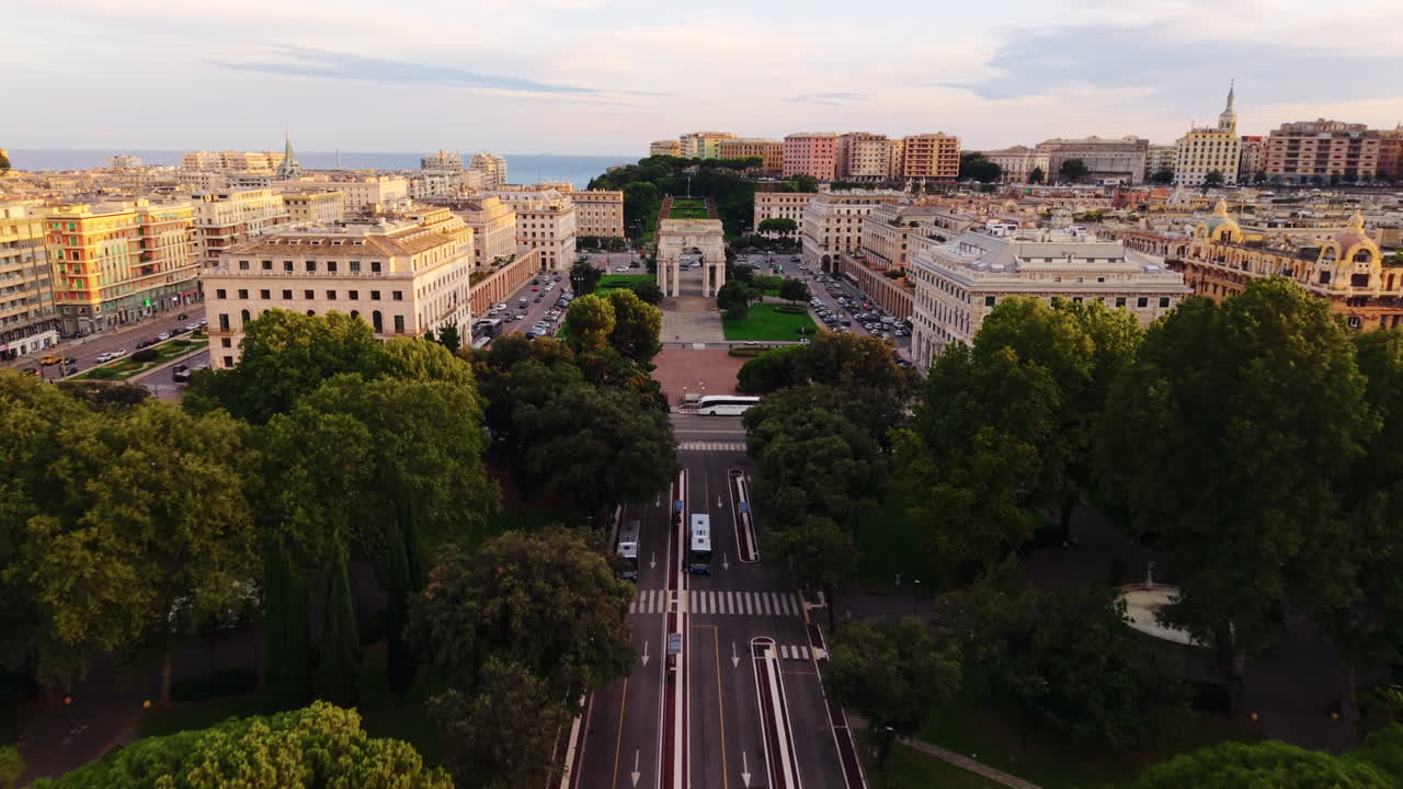 Drone moves forward above the tree-lined street towards Piazza della Vittoria with the arch, surrounding buildings, and sea view at sunset