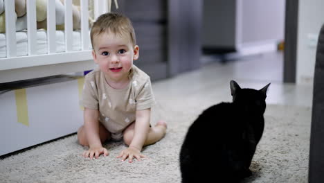 adorable boy sitting on the floor petting black cat. beautiful baby looks at camera and touches pet again. blurred backdrop.