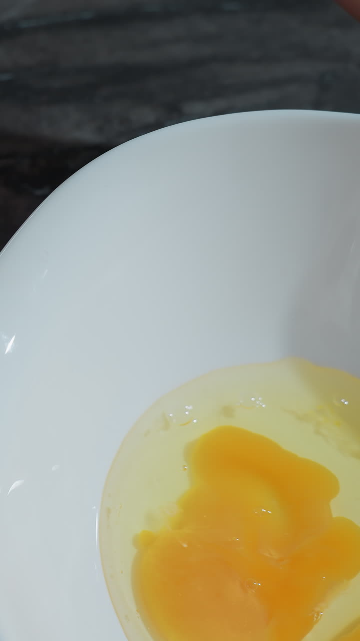 Close-up of a person hand preparing to crack an egg on a countertop, with a white bowl containing broken eggs nearby, the raw egg yolk and egg white are visible