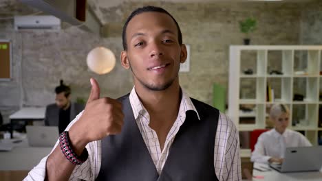 apuesto hombre de negocios africano está de pie y mirando a la cámara en la oficina, mostrando como señal, sonriendo, colegas están trabajando con computadoras portátiles, concepto de trabajo