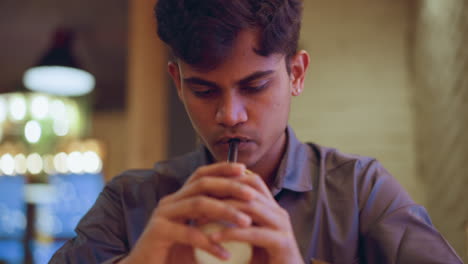Centered front view of young man drinking through black straw with eyes looking downward, hands clasped around cup, creating mood of quiet focus, introspection, or peaceful pause in warm indoor environment