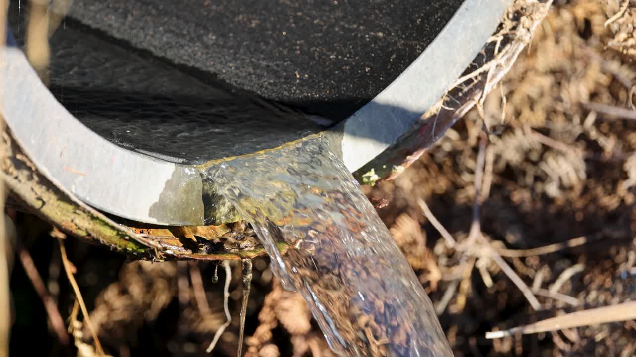 Clear water flows steadily from a pipe, surrounded by dry vegetation, under bright sunlight