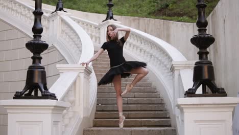 Front-view-of-a-graceful-ballet-movements.-Young,-blonde-haired-ballerina-doing-vertical-twine-and-posing-standing-on-a-beginning-of-street-stairs-in-black-tutu.-Daytime.-Slow-motion