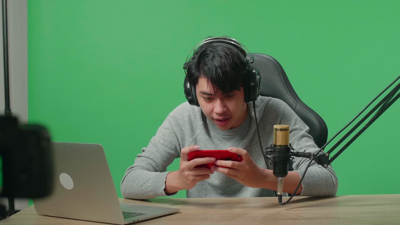 Asian Man Gamer With Headphone And Computer Celebrating Winning Mobile Phone Game While Shooting Video By Camera On Green Screen