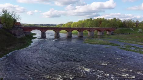 clip aéreo 4k, volando sobre la pequeña ciudad de kuldiga letonia, puente de ladrillo rojo sobre el río venta, amplia cascada