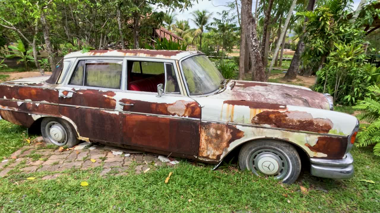 Weathered antique sedan sits abandoned on grass, surrounded by tropical plants, daylight, slow camera pan