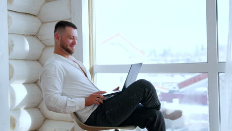 handsome caucasian male in white shirt sits at the window. man looks at laptop smiling pressing its key.