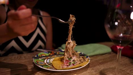 a woman in a chevron dress uses a fork to lift shredded pork from a taco on a colorful plate in a dimly lit restaurant