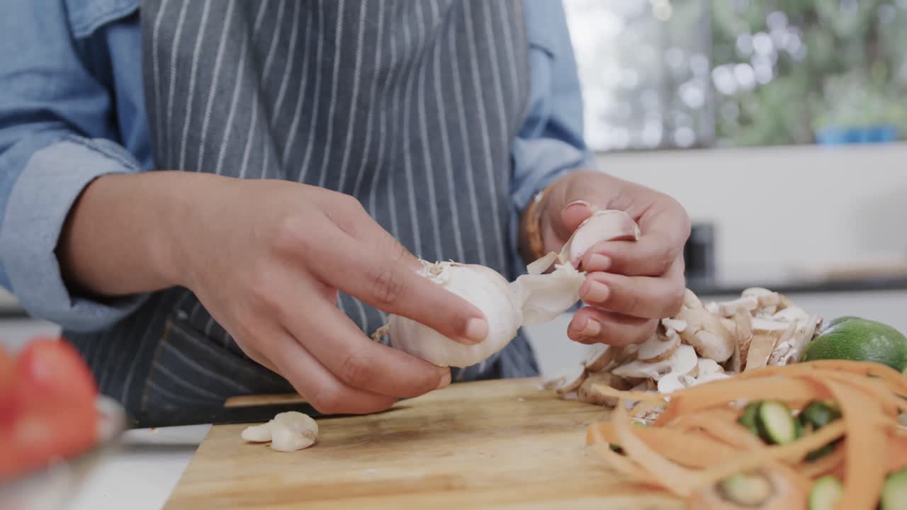 Midsection of biracial woman in apron preparing meal, peeling garlic in kitchen, slow motion