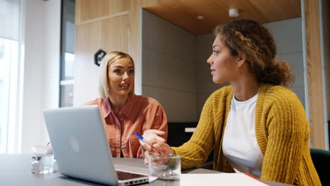 Low-angle-view-of-two-smiling-young-female-colleagues-sitting-in-an-office-working-together-at-a-laptop-computer,-close-up