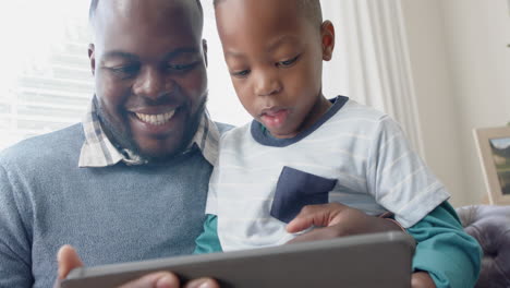 Feliz-Padre-Afroamericano-Sentado-En-El-Sofá-Con-Su-Hijo-En-El-Regazo-Usando-Una-Tableta,-En-Cámara-Lenta