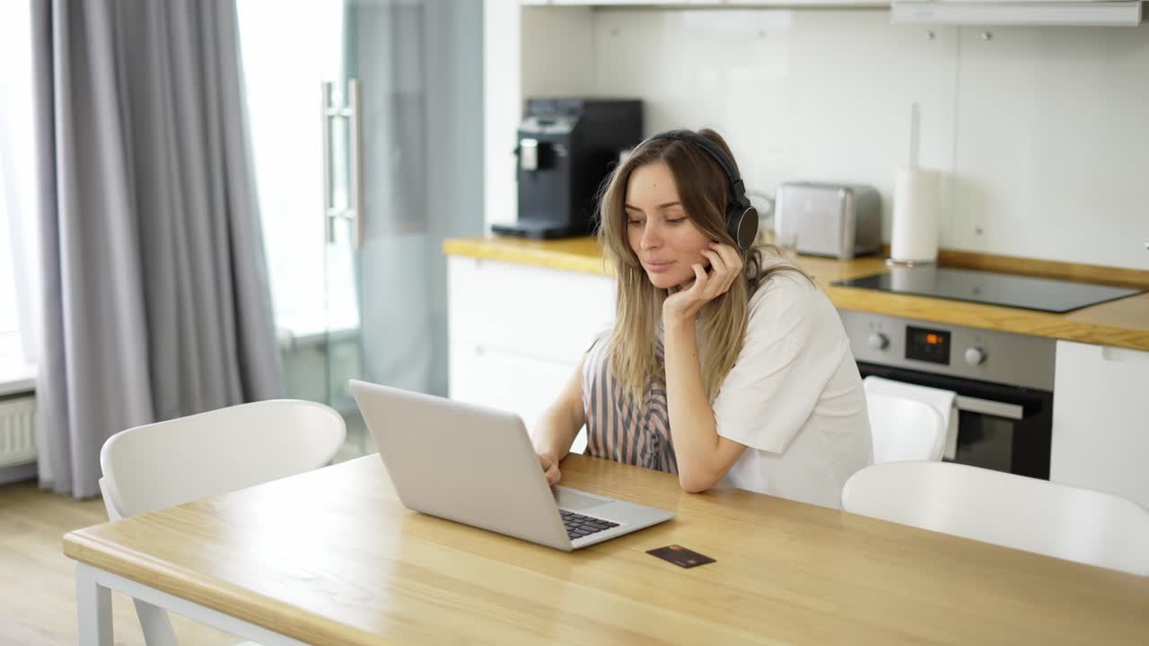 Donna con le cuffie in cucina, che ascolta musica e fa un acquisto online usando una carta di credito e un portatile.