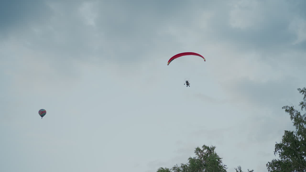 upward perspective showing powered paraglider under red canopy flying against dramatic cloudy sky alongside distant hot air balloon, scene captures serene aerial contrast
