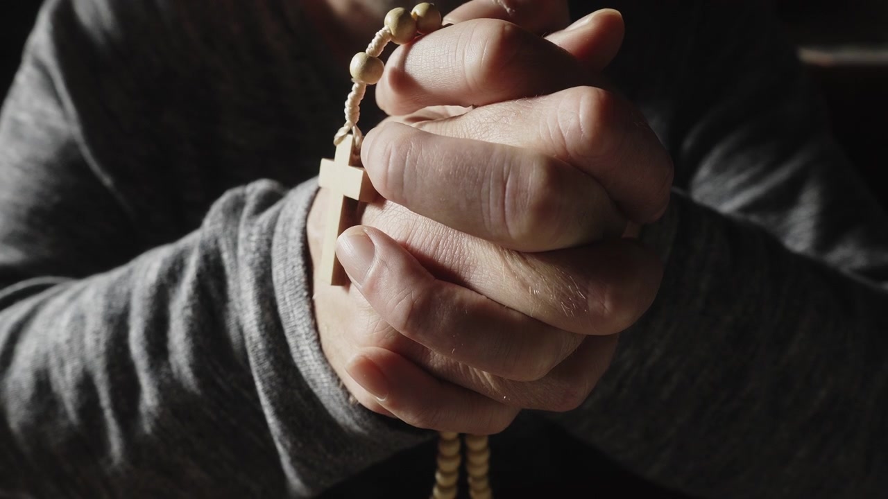 A dramatic close-up of tightly clasped hands holding a crucifix rosary in deep prayer and quiet meditation. Perfect for themes of faith, grief, hope, and spiritual reflection.