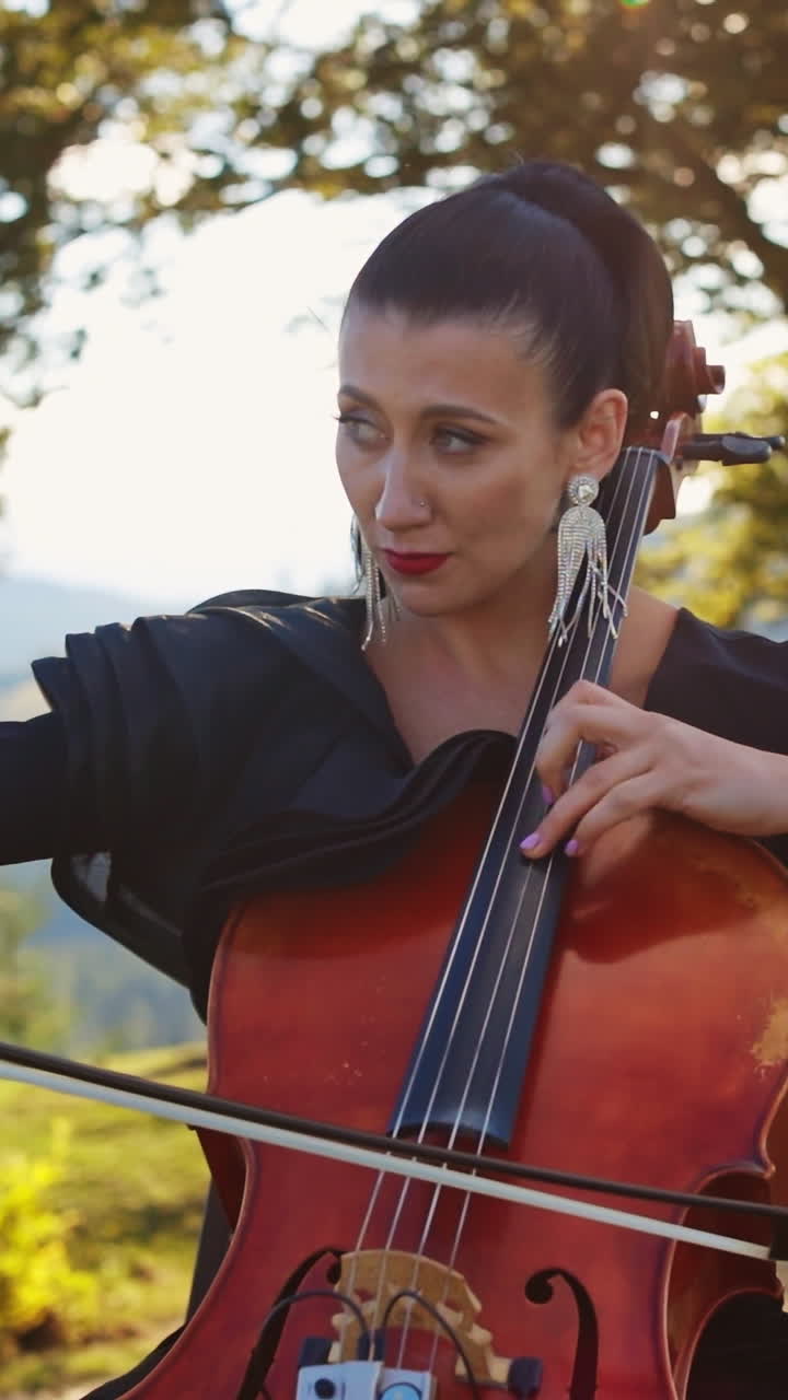 Violoncello musician with bright makeup and long earrings playing instrument. Lady stops to fix the earring and goes back to playing. Nature backdrop. Vertical video