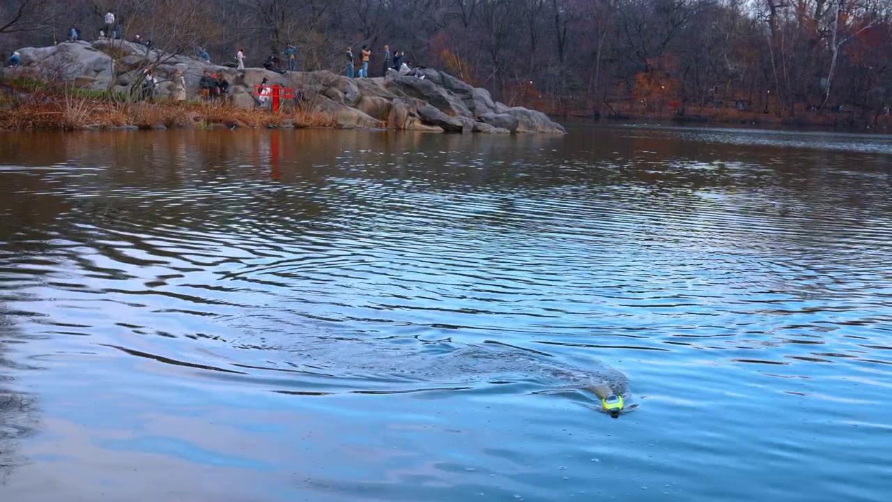 New York, USA, 24 March 2026: Remote control boat on lake in Central Park. A small yellow toy boat creating ripples on the water with people on rocks in the background.