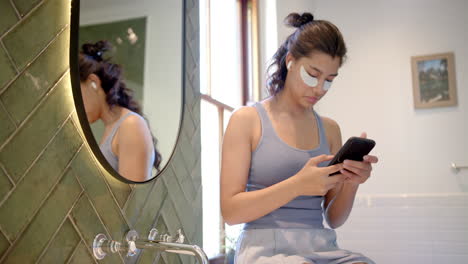 biracial teenage girl wearing under eye masks using smartphone in bathroom, copy space, slow motion