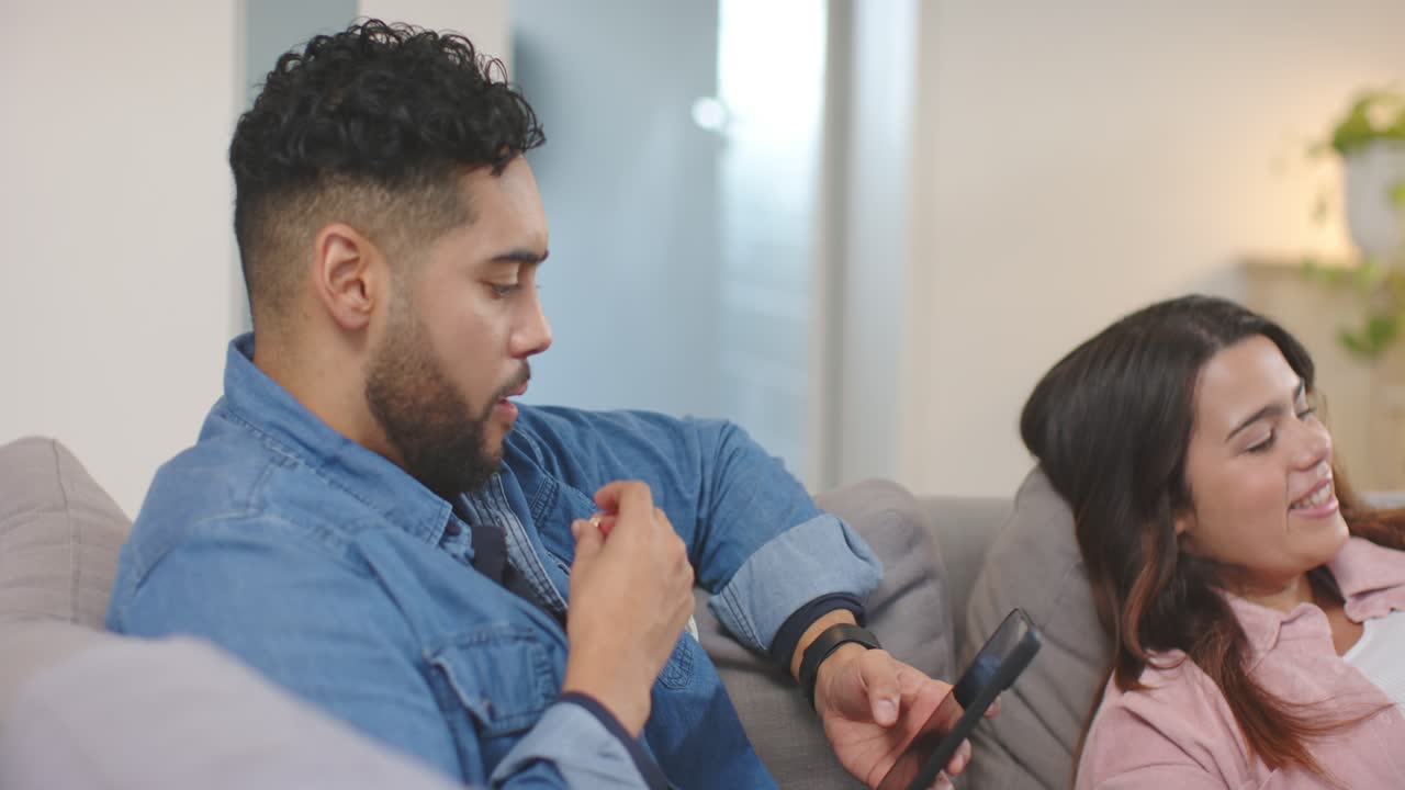 Coppia multiculturale annusa popcorn, l'uomo ne prende e sgranocchia mentre controlla lo smartphone sul divano.