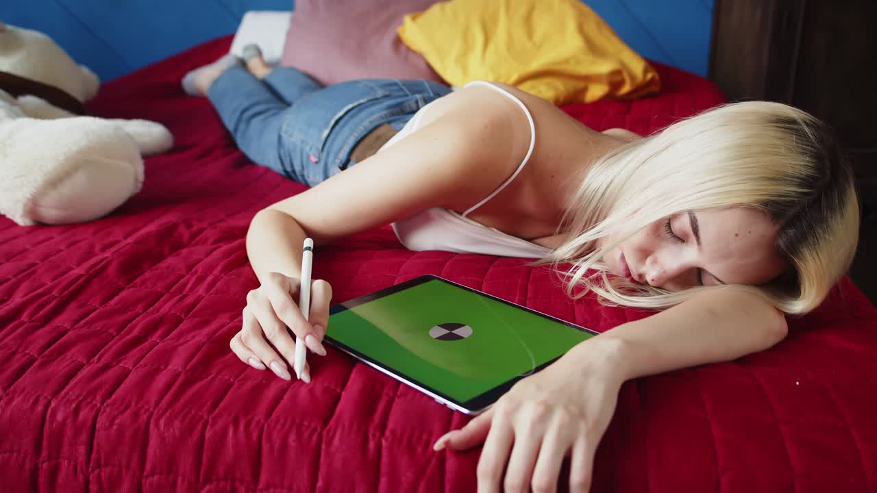 피곤한 금발 여성은 침대에 누워 잠을 자고, 그녀의 손에 스타일러스를 들고, 그 에는 녹색 화면, 모형, chromakey의 디지털 태블릿이 있습니다.