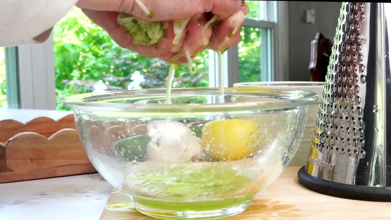 Squeezing water out of cucumbers. Making Tzatziki Greek or Mediterranean sauce or dip for a party. Dip or sauce in a green bowl on a wooden cutting board with dill, green napkin and a grater.