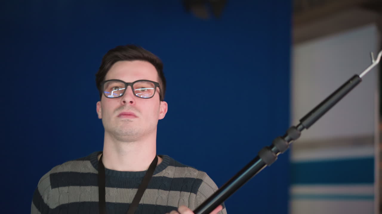 Close-up of man holding boom microphone in professional studio, staring directly at camera with focused expression, preparing for sound recording during video production, clear blue backdrop